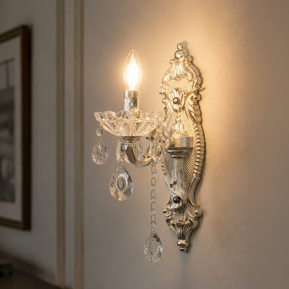 An ornate wall-mounted sconce with a warm gold-toned metal frame, a lit candle-style bulb, and hanging clear crystal prisms, shown mounted on a beige wall in an interior setting.