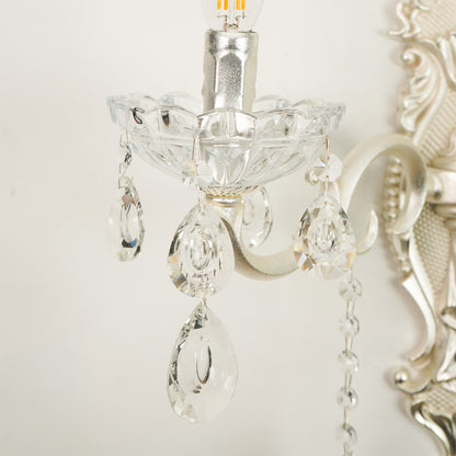 Close-up of a silver wall sconce with clear cut glass teardrop crystal drops and a visible light bulb, shown against a light neutral wall.