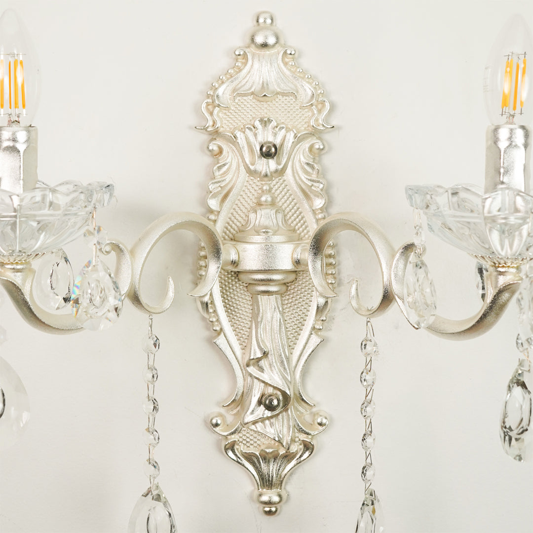 An ornate silver wall sconce with intricate baroque-style detailing, clear crystal candle-style holders, and hanging teardrop crystal pendants, shown mounted against a plain off-white wall.
