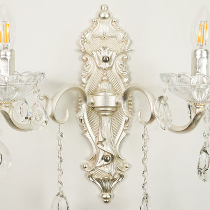 An ornate silver wall sconce with intricate baroque-style detailing, clear crystal candle-style holders, and hanging teardrop crystal pendants, shown mounted against a plain off-white wall.