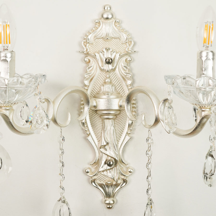 An ornate silver wall sconce with intricate baroque-style detailing, clear crystal candle-style holders, and hanging teardrop crystal pendants, shown mounted against a plain off-white wall.