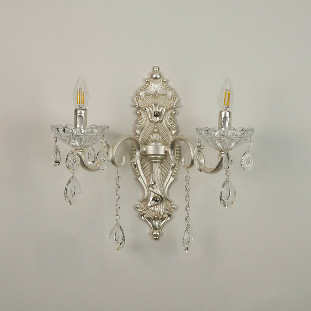 An ornate silver wall sconce with two candle-style lights, clear crystal teardrop pendants, and a decorative central crest, shown against a plain light gray wall.
