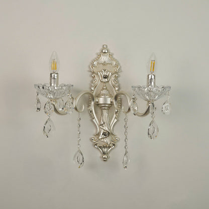 An ornate silver wall sconce with two candle-style lights, clear crystal teardrop pendants, and a decorative central crest, shown against a plain light gray wall.