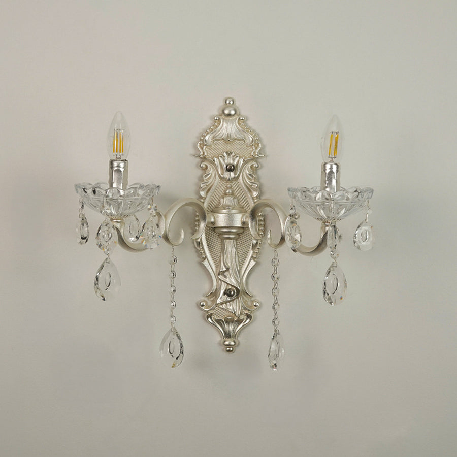 An ornate silver wall sconce with two candle-style lights, clear crystal teardrop pendants, and a decorative central crest, shown against a plain light gray wall.