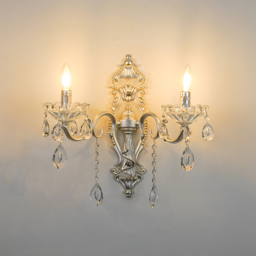 An ornate silver wall sconce with two lit candle-style bulbs and hanging clear crystal teardrop drops, mounted on a plain light gray wall.