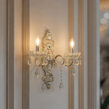 An ornate silver wall sconce with two lit candle-style bulbs and hanging clear crystal prisms, mounted on a beige paneled wall.