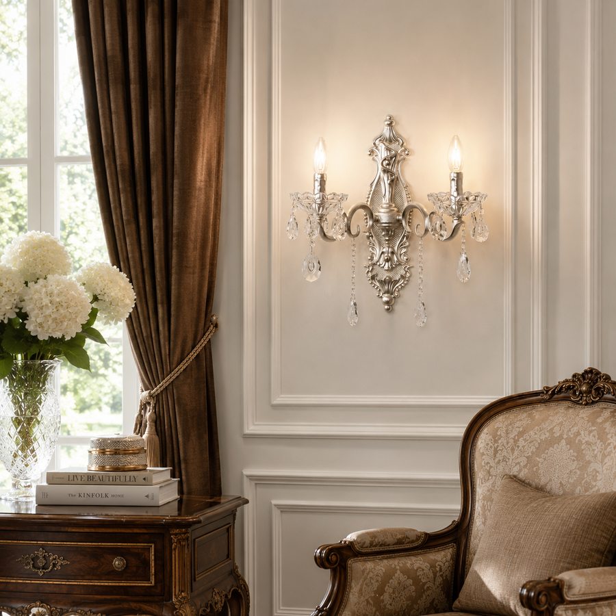An ornate silver wall sconce with crystal droplets and two lit candle-style bulbs, mounted on a cream paneled wall in a classic room setting with brown curtains, a wooden side table, and a vintage armchair.
