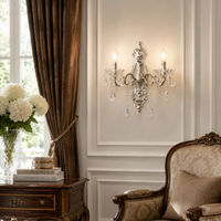 An ornate silver wall sconce with crystal droplets and two lit candle-style bulbs, mounted on a cream paneled wall in a classic room setting with brown curtains, a wooden side table, and a vintage armchair.