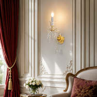 An ornate gold and crystal wall sconce mounted on a cream paneled wall in a luxurious room with red drapery, a floral arrangement, and part of a classic armchair.