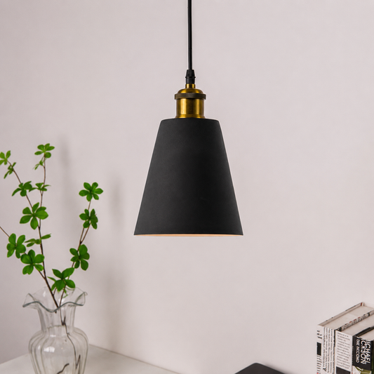 A modern pendant light with a matte black cone-shaped shade, a brass-finished metal collar, and a black hanging cord, shown against a light wall above a white surface with a small vase of green stems and books nearby.