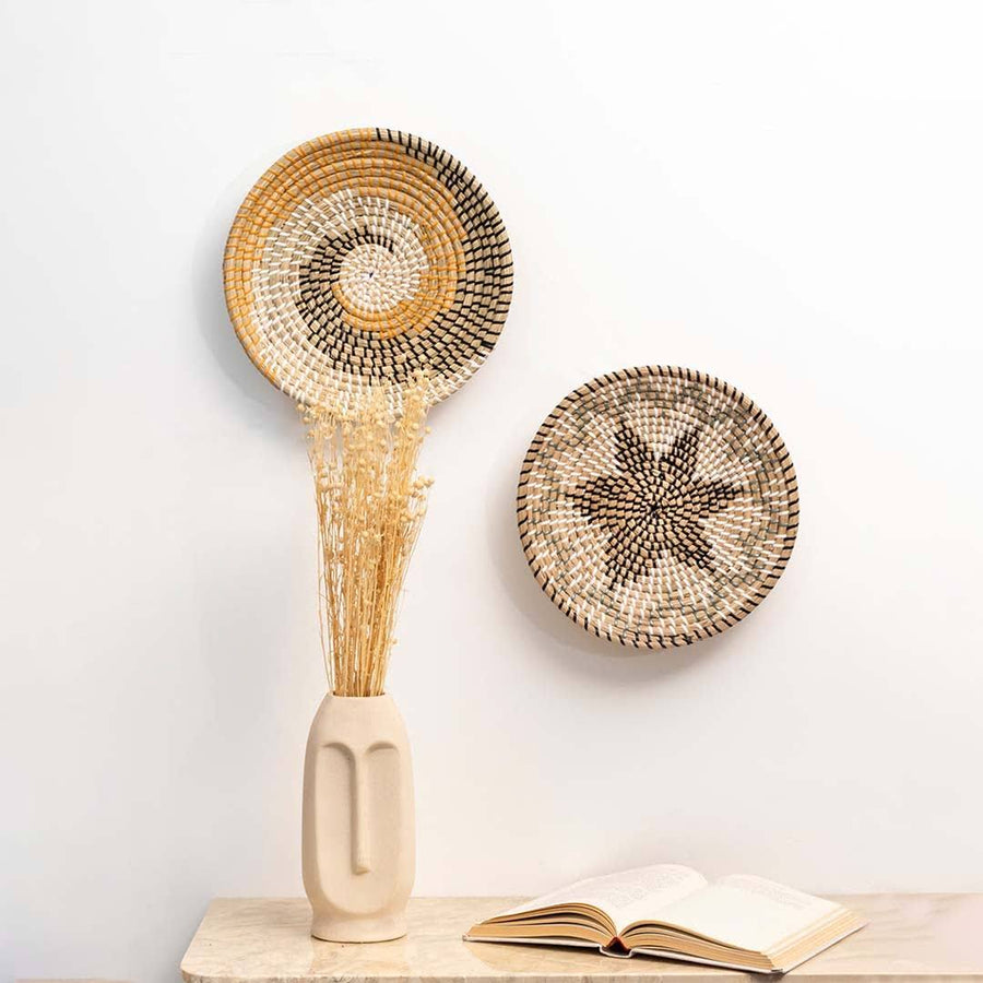 Two woven seagrass wall baskets styled above a table console setup featuring a ceramic face vase and an open book.