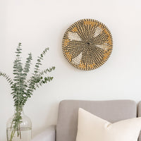 A round woven wall basket with a black and beige spiral pattern hangs on a white wall above a grey sofa beside a vase of green eucalyptus branches.