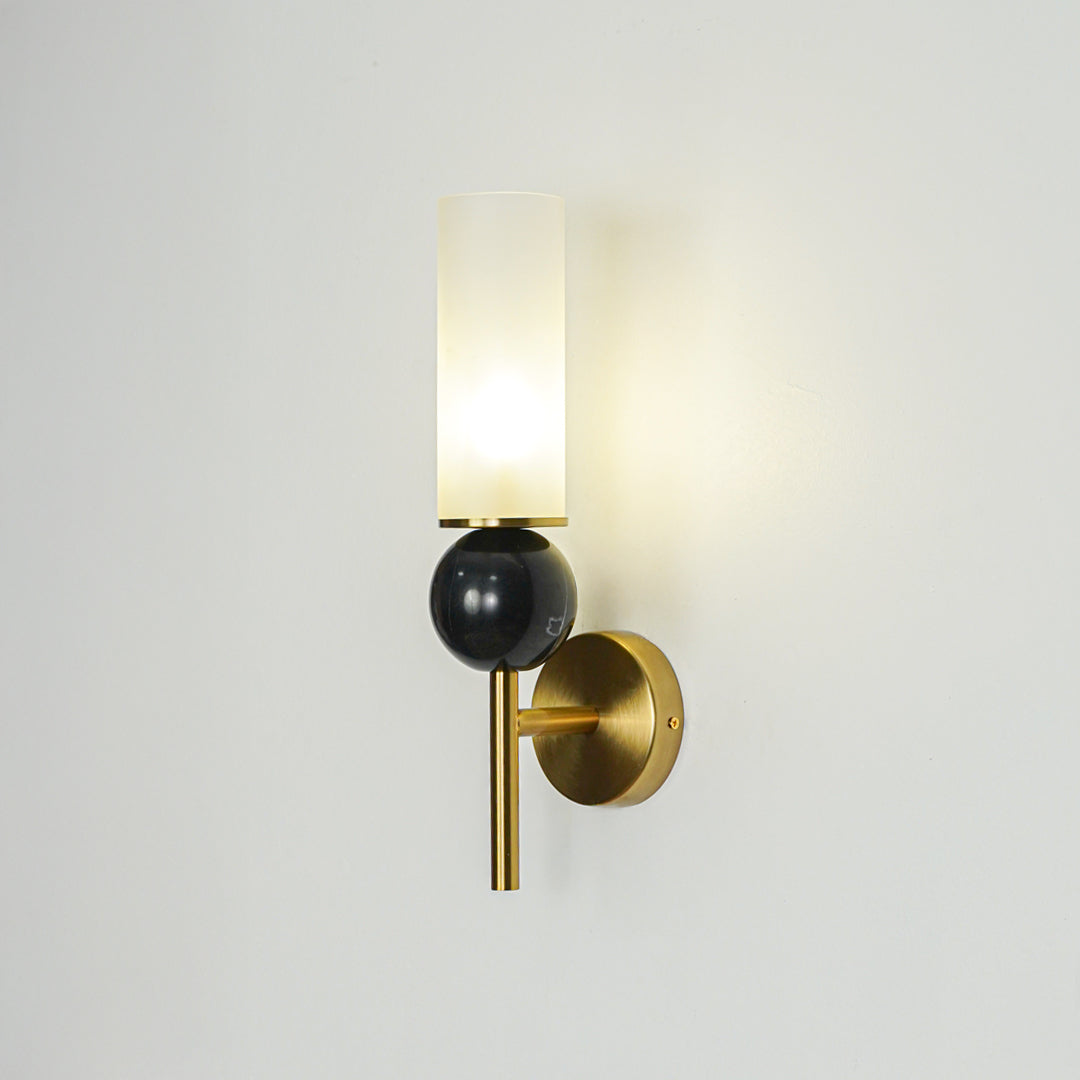 Contemporary black and gold wall light with frosted glass cylinder mounted on neutral wall background creating warm glow for modern interiors.