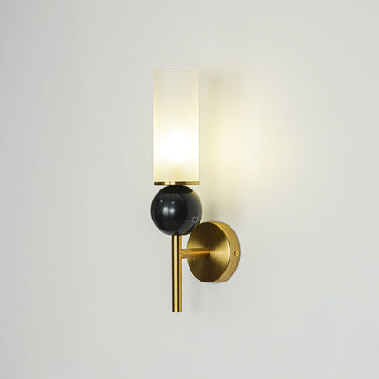 Contemporary black and gold wall light with frosted glass cylinder mounted on neutral wall background creating warm glow for modern interiors.