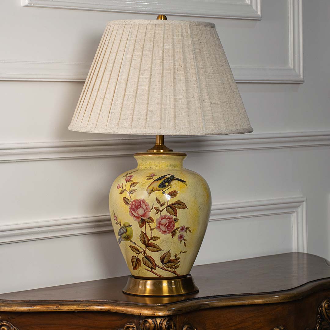 Yellow ceramic floral table lamp with beige fabric shade displayed on white wall background with lights off.
