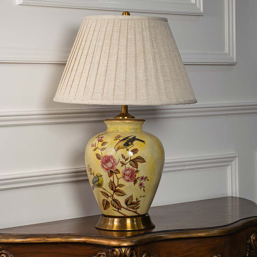 Yellow ceramic floral table lamp with beige fabric shade displayed on white wall background with lights off.