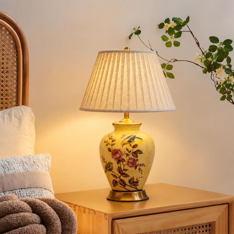 A decorative table lamp with a pleated beige shade and a yellow ceramic base decorated with pink flowers and green leaves, placed on a wooden bedside table in a warm, cozy room setting.