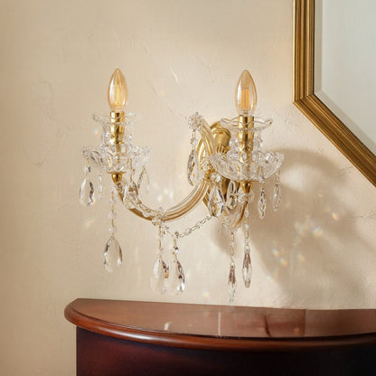 A wall-mounted two-light sconce with a polished gold-tone metal frame and clear crystal droplets, shown installed above a dark wood console table against a beige wall.