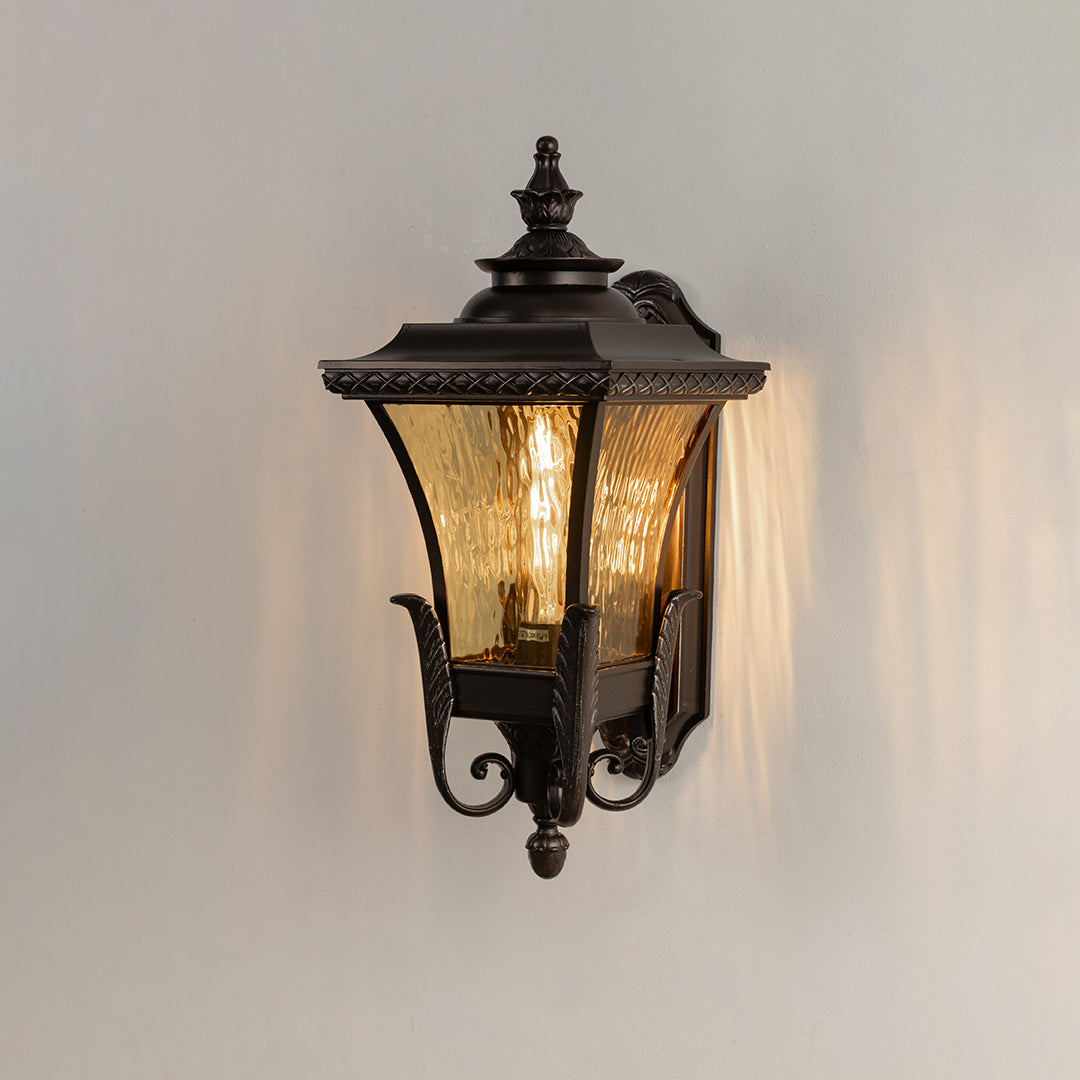 Classic bronze metal exterior sconce with amber ripple glass glowing on white background for patio wall placement.