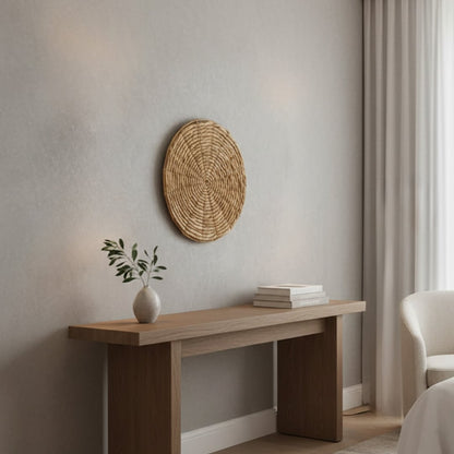Lifestyle image of round woven wall decor mounted on a neutral wall above a wooden console table styled with minimal decor in a calm modern living space.