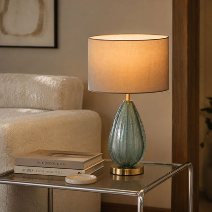 A table lamp with a textured, ribbed teal-green glass base and a warm beige fabric drum shade, lit up on a glass-topped side table in a cozy living room setting.