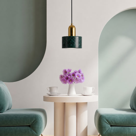 A modern pendant light with a textured dark green drum shade and a gold neck hangs above a round cream table holding a vase of purple flowers, flanked by two teal armchairs in a minimalist interior setting.