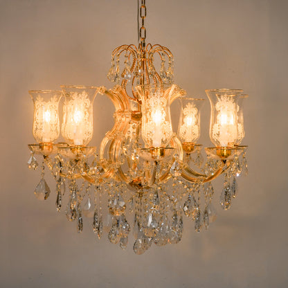 A classic six-light crystal chandelier with a gold-finished metal frame, illuminated glass shades, and hanging clear crystal prisms, shown against a plain beige wall.