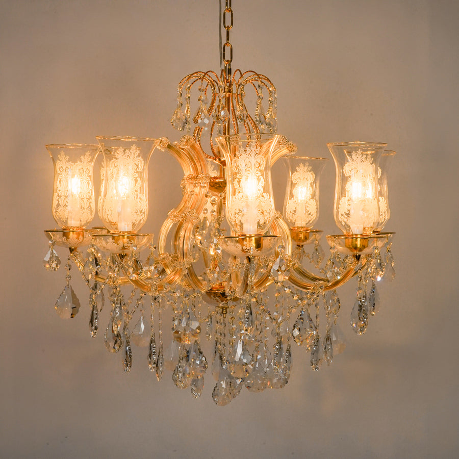 A classic six-light crystal chandelier with a gold-finished metal frame, illuminated glass shades, and hanging clear crystal prisms, shown against a plain beige wall.