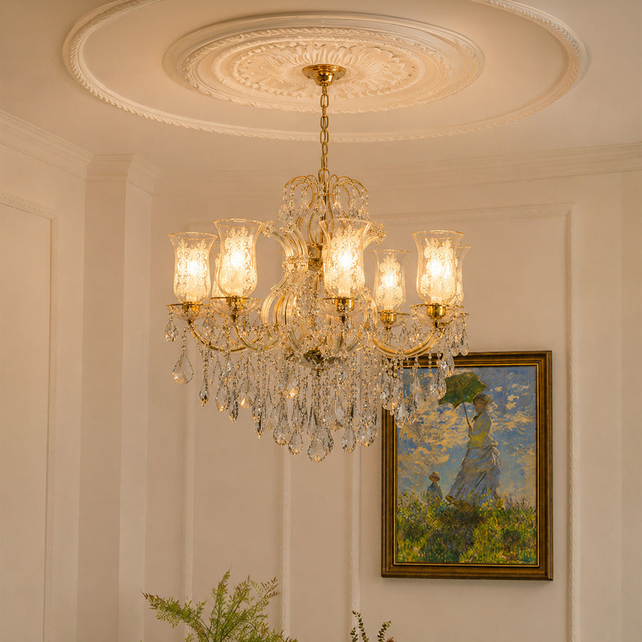 A multi-light crystal chandelier with gold-toned metal arms and hanging clear glass prisms, suspended from a decorative cream-colored ceiling medallion in an elegant room. A framed landscape painting is visible on the wall behind it.