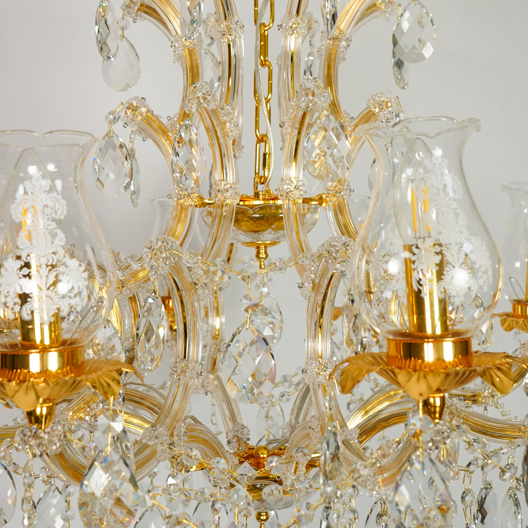 Close-up of an ornate chandelier with gold-finished metal arms and clear glass shades and hanging crystal prisms against a light neutral background.
