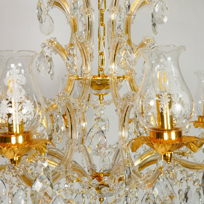 Close-up of an ornate chandelier with gold-finished metal arms and clear glass shades and hanging crystal prisms against a light neutral background.