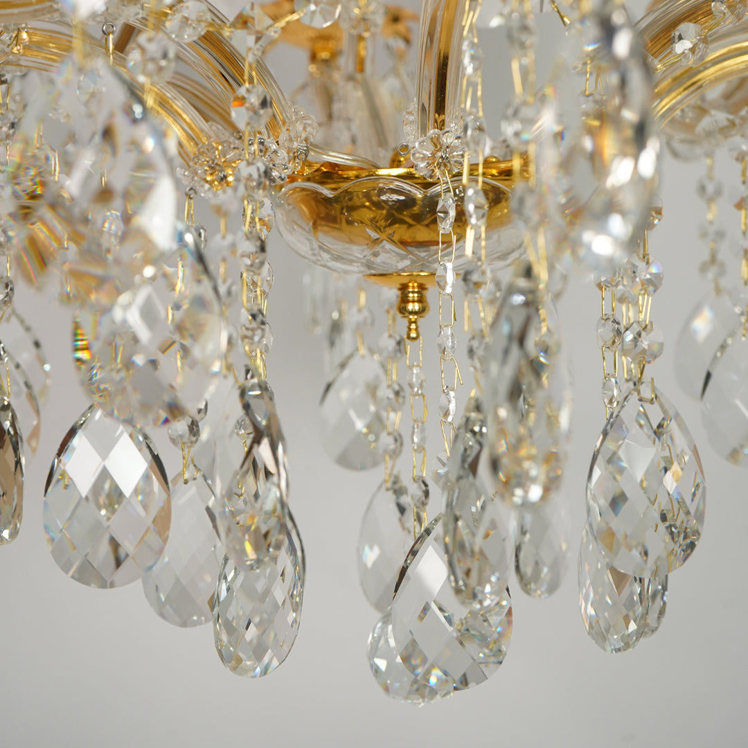 Close-up of a crystal chandelier with faceted clear teardrop pendants and gold-toned metal arms against a light gray background.