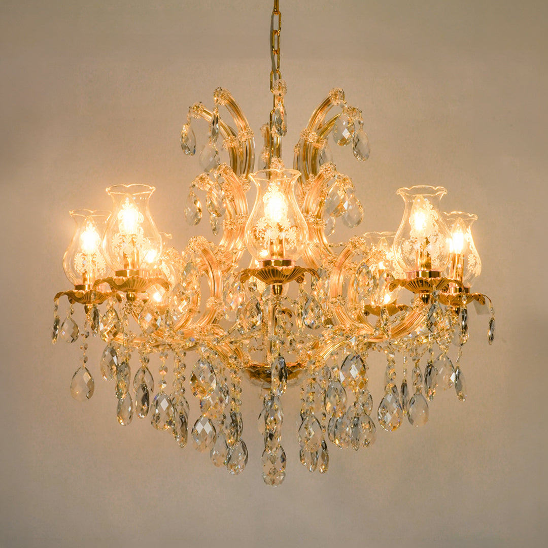 A multi-light crystal chandelier with a gold-tone metal frame and hanging clear glass teardrop prisms, shown illuminated against a plain beige wall.