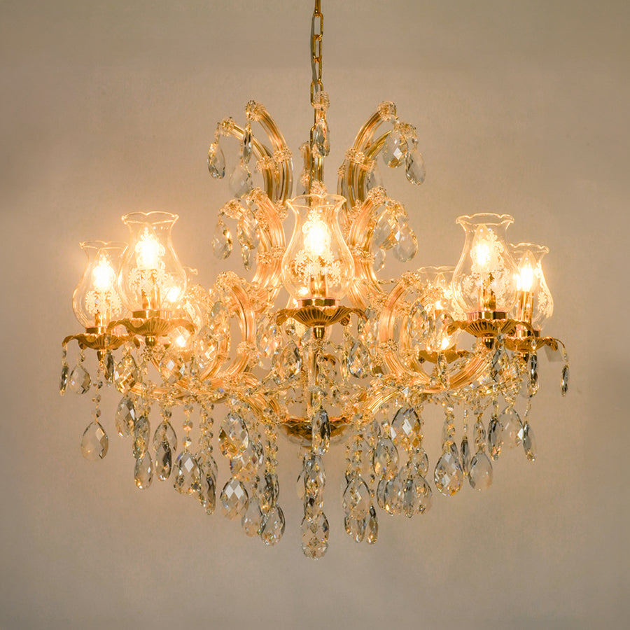 A multi-light crystal chandelier with a gold-tone metal frame and hanging clear glass teardrop prisms, shown illuminated against a plain beige wall.