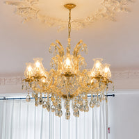 An ornate gold chandelier with multiple clear crystal hanging drops and bell-shaped glass shades, illuminated with warm light, hanging from a decorative ceiling in a room with white curtains.