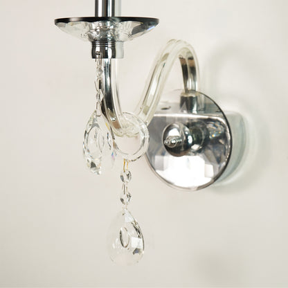 A close-up of a wall-mounted light fixture with a polished silver metal arm and circular wall plate, adorned with clear hanging crystal prisms against a plain off-white wall.