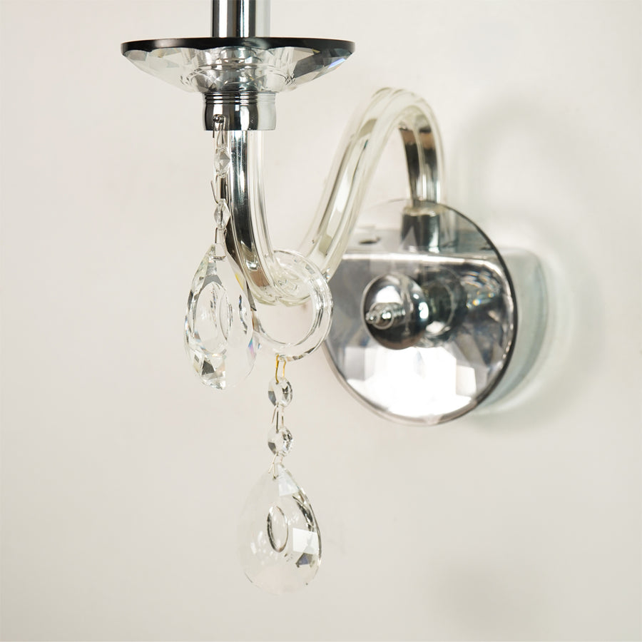A close-up of a wall-mounted light fixture with a polished silver metal arm and circular wall plate, adorned with clear hanging crystal prisms against a plain off-white wall.