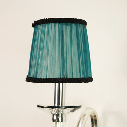 A teal pleated fabric lampshade with black braided trim at the top and bottom, mounted on a chrome metal lamp stem against a plain off-white background.
