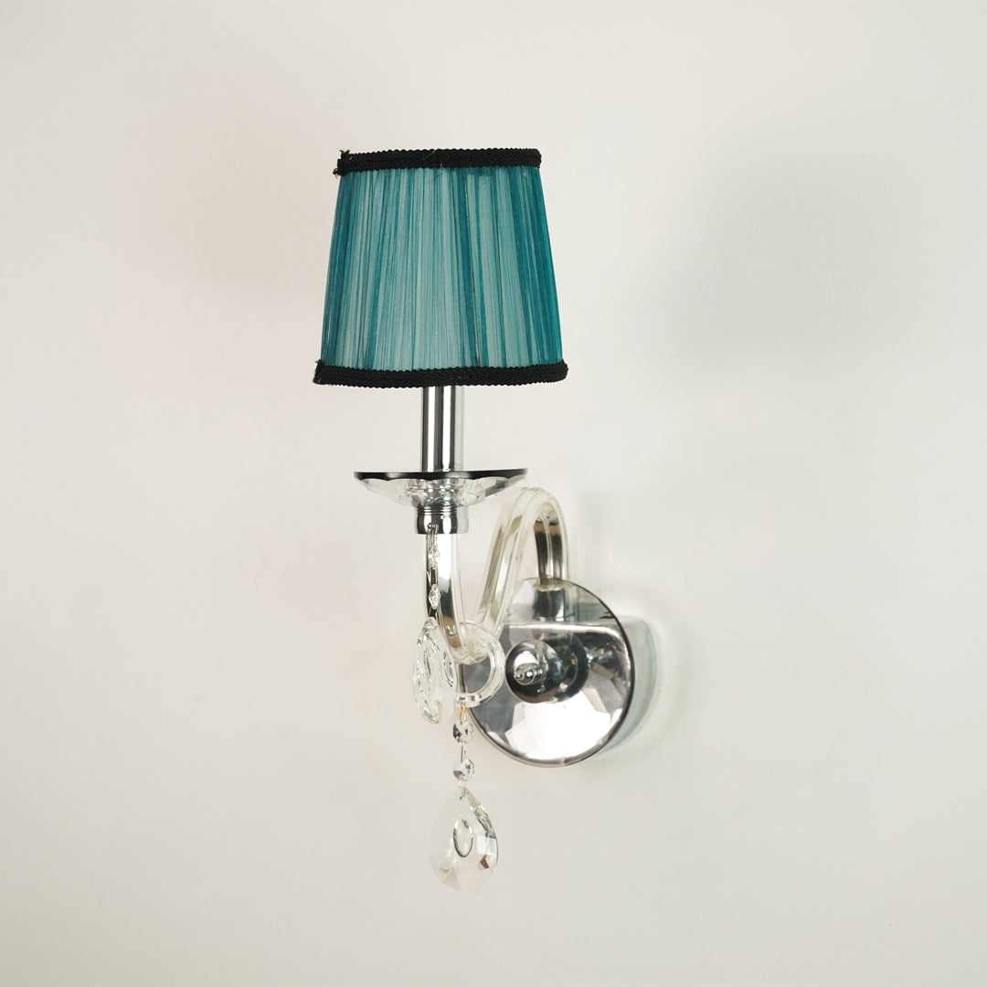 A wall-mounted sconce with a teal pleated fabric shade trimmed in black, mounted on a polished silver curved arm with clear hanging crystal drops against a plain off-white wall.