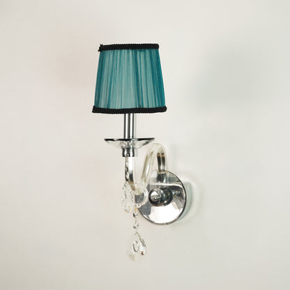 A wall-mounted sconce with a teal pleated fabric shade trimmed in black, mounted on a polished silver curved arm with clear hanging crystal drops against a plain off-white wall.