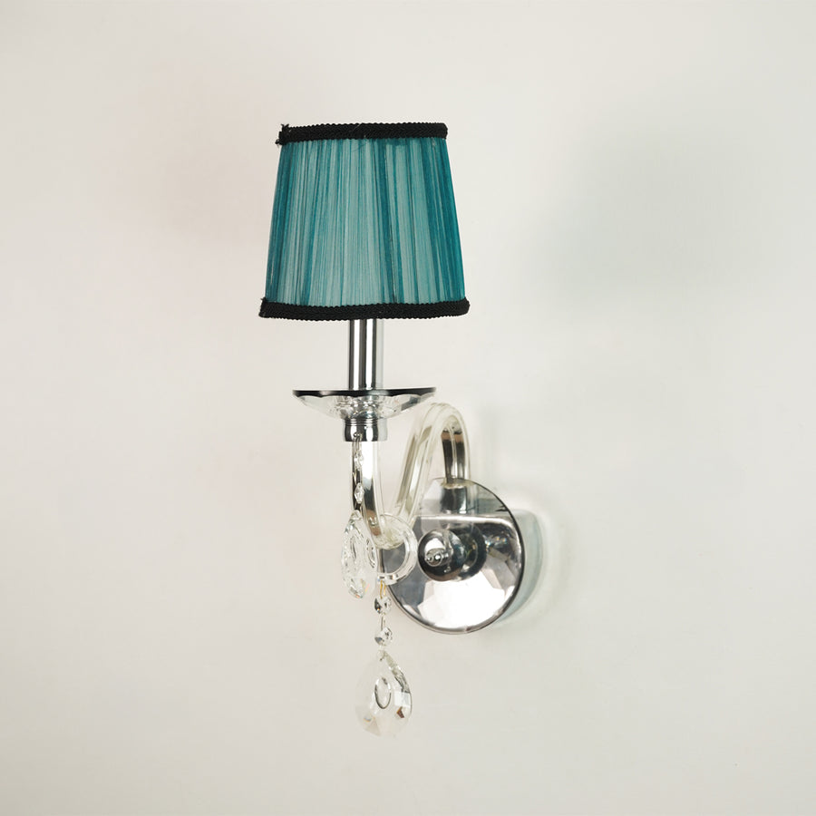 A wall-mounted sconce with a teal pleated fabric shade trimmed in black, mounted on a polished silver curved arm with clear hanging crystal drops against a plain off-white wall.
