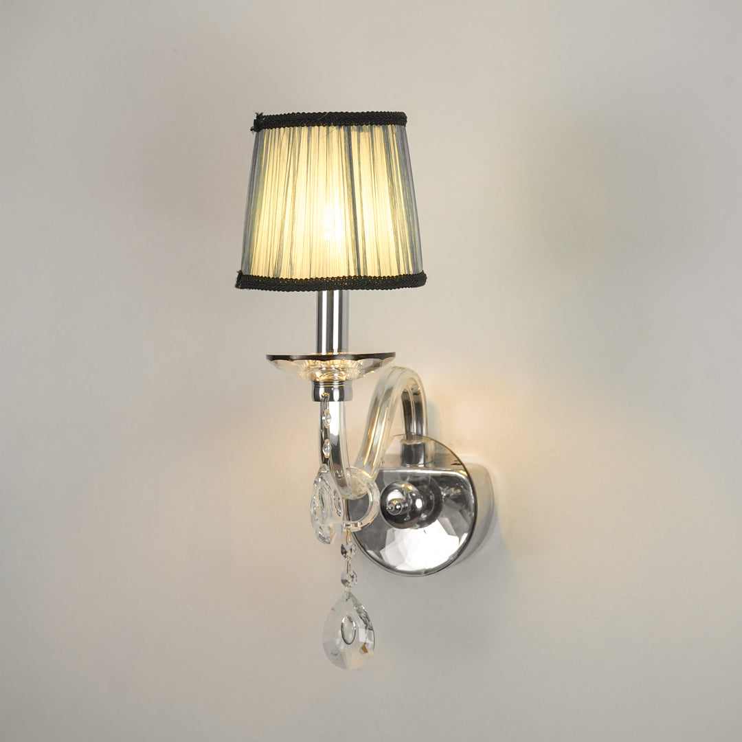 A wall-mounted sconce with a pleated beige fabric shade, black trim, a polished silver metal arm and wall mount, and clear hanging crystal teardrop ornaments, shown lit against a plain light wall.