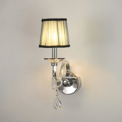 A wall-mounted sconce with a pleated beige fabric shade, black trim, a polished silver metal arm and wall mount, and clear hanging crystal teardrop ornaments, shown lit against a plain light wall.