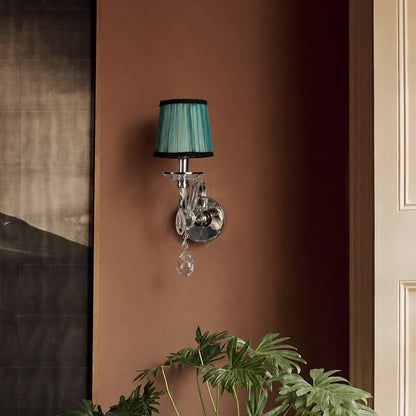 A wall-mounted sconce with a pleated teal green lampshade, polished silver metal body, and hanging clear crystal teardrop, shown installed on a warm brown wall with houseplant leaves at the bottom.