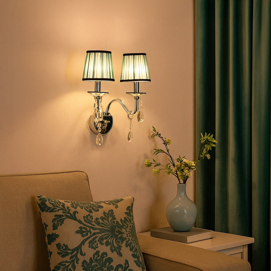 Double arm wall light with chrome base blue fabric shades and crystal drops glowing beside sofa and vase.
