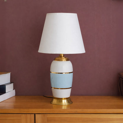 Table lamp with white and gold ceramic base and white shade on wooden table warm ambient lighting for living room decor.

