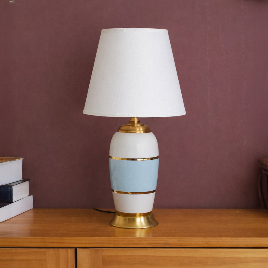 Table lamp with white and gold ceramic base and white shade on wooden table warm ambient lighting for living room decor.

