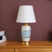 Table lamp with white and gold ceramic base and white shade on wooden table warm ambient lighting for living room decor.

