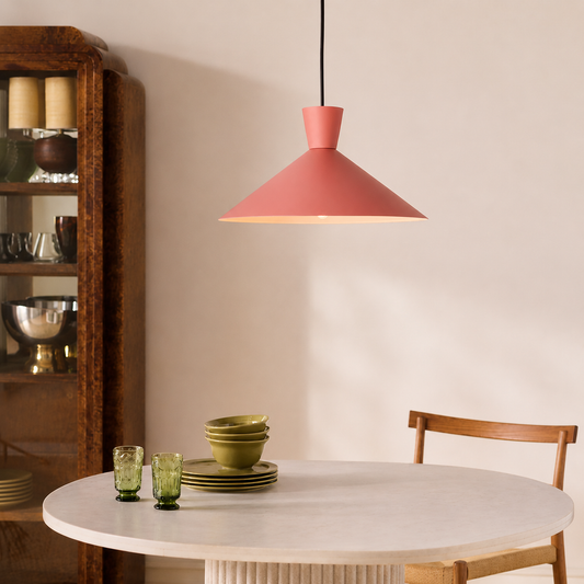 A modern pendant light with a dusty rose pink conical shade hangs over a round white dining table in a warm, styled room. A wooden chair is visible on the right, and a dark wood hutch stands on the left.