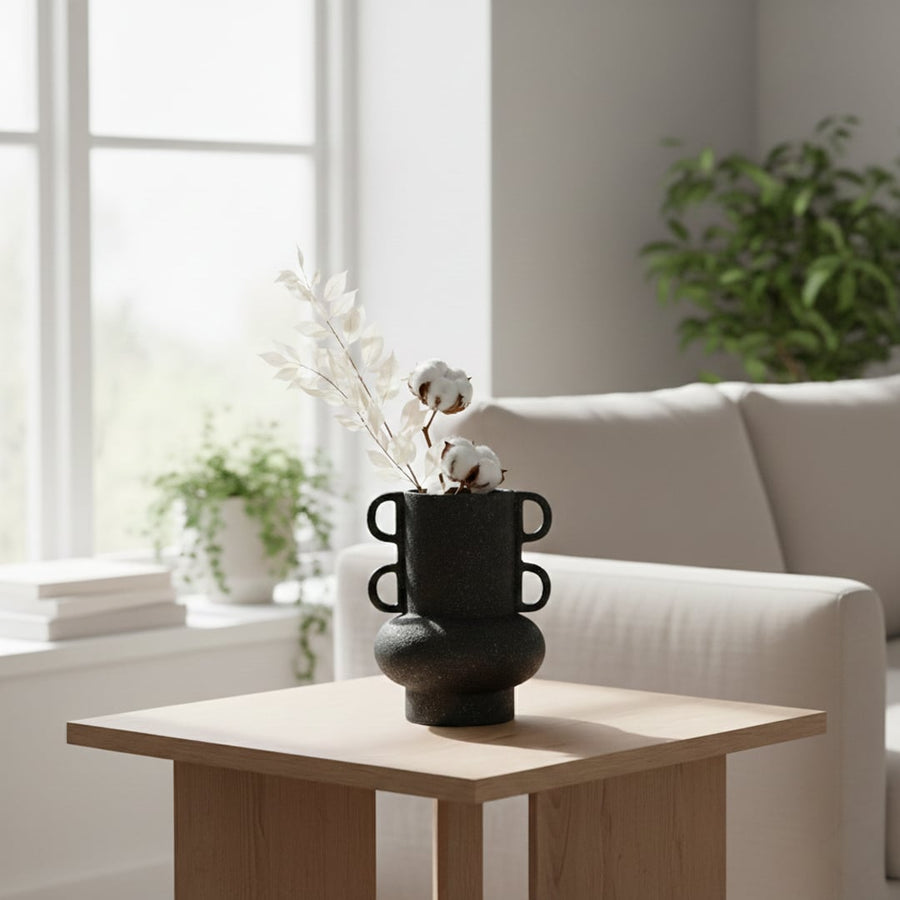 Black textured ceramic decorative vase styled with dried stems on wooden side table in bright modern living room setting.